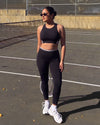 Woman in sunglasses standing on a tennis court with a racket, wearing QivoFit Black High-Rise Leggings with no front seam and grey contrast panels, with a matching sports bra.