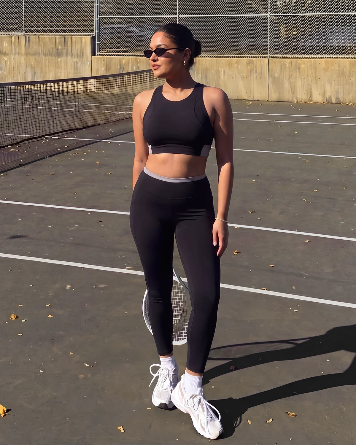 Woman in sunglasses standing on a tennis court with a racket, wearing QivoFit Black High-Rise Leggings with no front seam and grey contrast panels, with a matching sports bra.