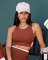 Woman in the QivoFit Brown Full Coverage Sports Bra with cream contrast panels and a white cap, seated at a café holding an iced coffee.