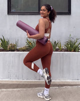 Smiling woman looking over her shoulder with a rolled yoga mat under her arm, wearing QivoFit Brown High-Rise Leggings and a matching full coverage racerback sports bra with cream contrast panels, one knee raised.