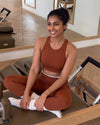 Smiling woman seated on a pilates reformer, wearing the QivoFit Brown Studio Set: Full Coverage Sports Bra and High-Rise Leggings with cream contrast panels.