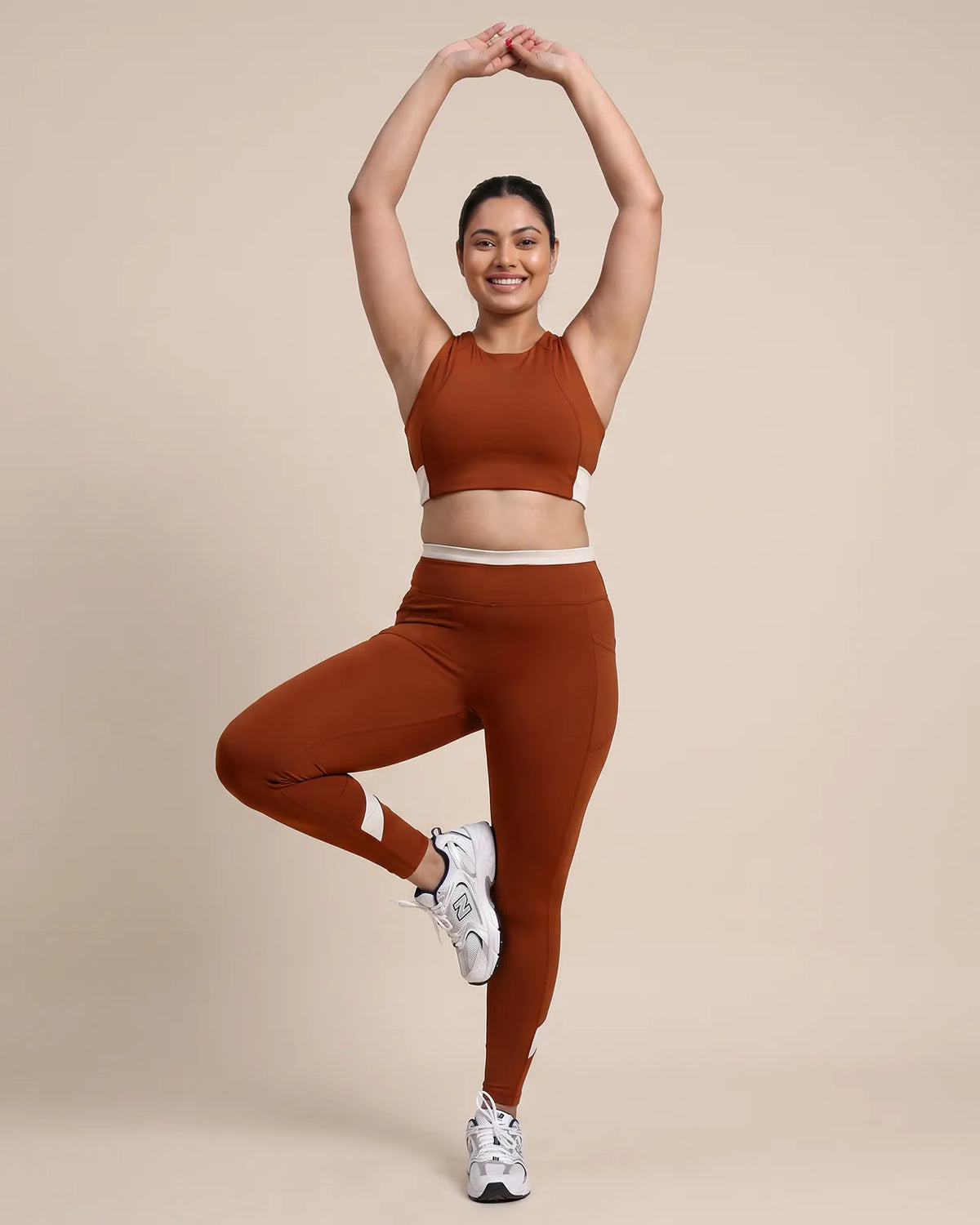 Full-length view of a model in a yoga tree pose, arms raised overhead, wearing the Brown Full Coverage Sports Bra and High-Rise Leggings.