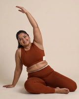 Smiling woman seated on the floor in a side stretch, one arm reaching overhead, wearing the Brown Full Coverage Sports Bra and High-Rise Leggings.