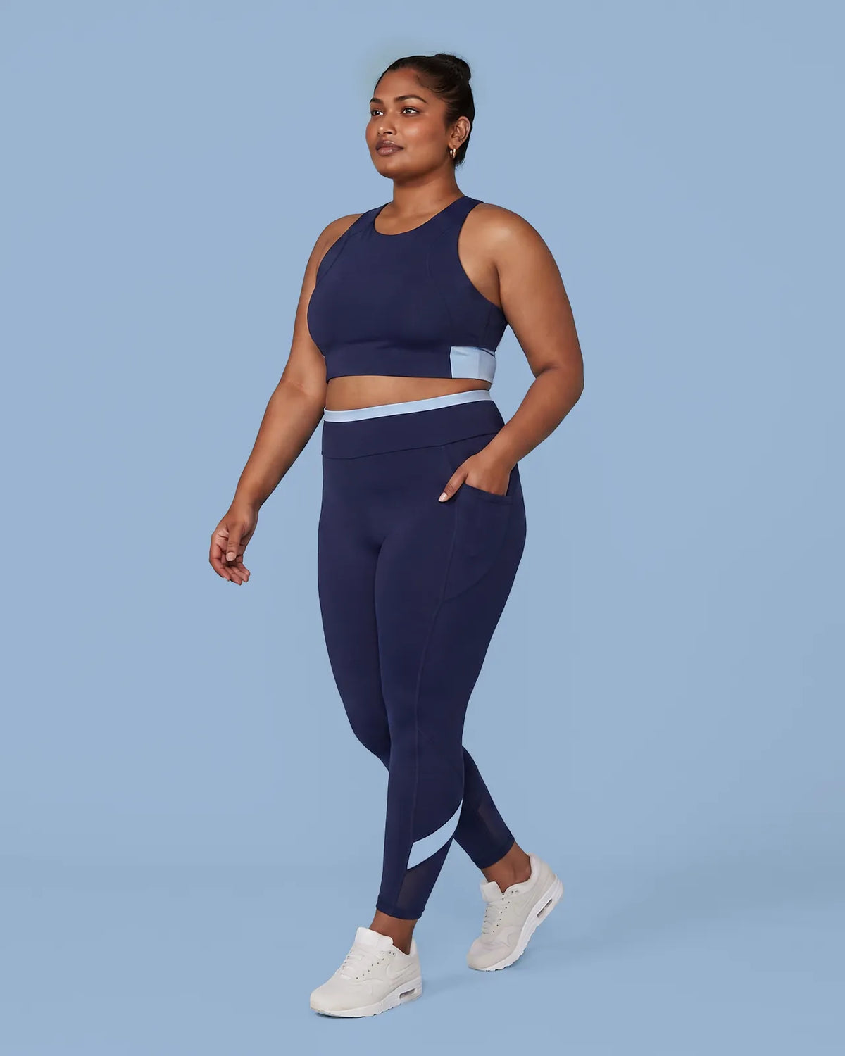 Woman walking in the Navy Full Coverage Sports Bra and High-Rise Leggings, one hand tucked into the side pocket.