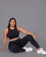 Woman seated on the floor leaning back on one hand, wearing the Black Full Coverage Sports Bra and High-Rise Leggings with white sneakers.