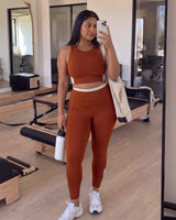 Woman taking a mirror selfie in a pilates studio with a tote bag and water bottle, wearing QivoFit Brown High-Rise Leggings with no front seam and a matching sports bra.