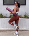 Smiling woman looking over her shoulder with a rolled yoga mat under her arm, wearing QivoFit Brown High-Rise Leggings and a matching full coverage racerback sports bra with cream contrast panels, one knee raised.