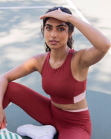 Woman in the QivoFit Maroon Full Coverage Sports Bra and matching leggings, seated on an outdoor court with one hand resting on her forehead.