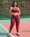 Smiling woman standing on an outdoor court in QivoFit Maroon High-Rise Leggings with a smooth front and blush pink calf panels, worn with a matching sports bra.
