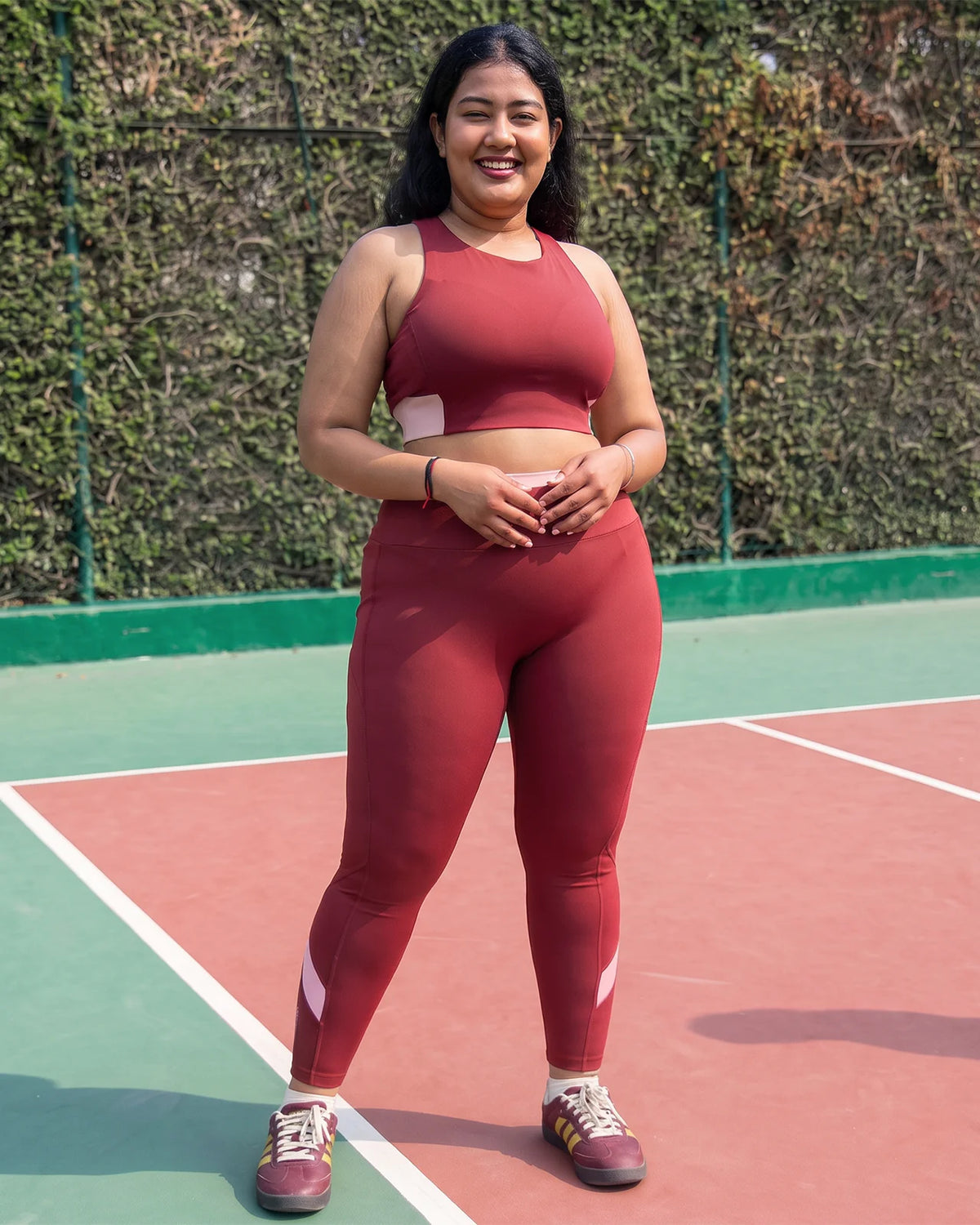 Smiling woman standing on an outdoor court in QivoFit Maroon High-Rise Leggings with a smooth front and blush pink calf panels, worn with a matching sports bra.