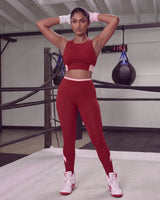 Woman standing in a boxing ring with hands behind her head, wearing the QivoFit Maroon Studio Set: Full Coverage Sports Bra and High-Rise Leggings with blush pink contrast panels.