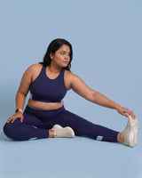 Woman seated on the floor reaching toward her foot in a hamstring stretch, wearing Navy High-Rise Leggings and a matching sports bra.