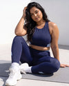 Woman seated on a mat in sunlight, one hand in her hair, wearing the QivoFit Navy Studio Set: Full Coverage Sports Bra and High-Rise Leggings with light blue contrast panels.